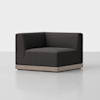 A modern, dark gray, L-shaped corner chair with a low wooden base sits on a light gray floor against a plain white background.