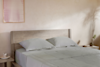 A neatly made bed with light-colored linens and two pillows sits against a beige wall. A small bedside table holds a cup, saucer, and a vase with greenery. A framed artwork hangs on the wall above the bed.