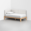 A minimalist daybed with a light wood frame, white mattress, white pillow, and cushioned white backrests is placed in the corner of a room with white walls and a gray floor.