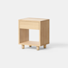 A small, light wood nightstand with a single drawer and an open shelf below, standing on four short cylindrical legs against a white background.