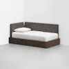 A modern daybed with a dark wooden frame and gray upholstered backrests is placed in the corner of a minimalist room. The bed is made with a white sheet and pillow.