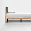 A minimalist wooden bed frame with a padded gray headboard, white sheets, and a pillow, placed against a plain white wall on a smooth white floor.