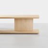 A minimalist light wood coffee table with clean lines and a central vertical support, placed on a light gray floor against a white background.