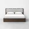 A modern bed with a dark wooden frame, a light gray upholstered headboard, and white bedding with two pillows, set against a plain white wall and light gray floor.