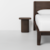 A modern, dark wood nightstand is placed next to a matching wooden bed frame with white bedding in a minimal, bright room with a white wall and floor.
