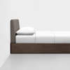 Side view of a neatly made bed with a brown wooden frame, a gray upholstered headboard, white sheets, and a white pillow against a plain white wall and floor.