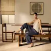 A woman in a beige tank top and dark pants sits on a wooden bench in a modern dining room, with a table, chairs, and minimalist wall art in the background.