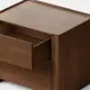 A simple, modern wooden nightstand with a natural finish, featuring one drawer partially open. The background is plain and light, highlighting the clean lines and minimalist design of the nightstand.