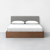 A modern bed with a gray upholstered headboard, white bedding, two white pillows, and a wooden frame, set against a plain white wall on a light gray floor.