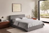 A modern bedroom with a gray upholstered bed, light gray bedding, two wooden nightstands, a table lamp, a potted plant, a framed abstract painting, and large glass doors opening to a garden.