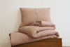 A folded blush pink quilt, matching pillow, and sheet set are neatly stacked on a wooden bench against a textured white wall.