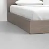 A close-up view of the corner of a modern wooden bed frame with a white mattress, sheets, and pillow, placed on a smooth light gray floor next to a white wall.