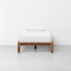 A single bed with a wooden frame, white bedding, and a white pillow, placed against a plain white wall on a light gray floor.