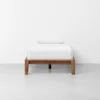 A single bed with a wooden frame, white bedding, and a white pillow, placed against a plain white wall on a light gray floor.