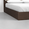 A close-up of a modern bed with a dark wooden frame, white sheets, and two white pillows, placed on a light gray floor against a plain white wall.