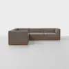 A modern L-shaped sectional sofa with brown upholstery and a wooden base sits in a minimal, light-colored room with a plain white background.