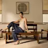 A woman in a sleeveless beige top and dark pants sits on a wooden bench in a minimalist dining room with matching table and chairs, soft lighting, and neutral-toned decor.