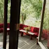 A cozy outdoor deck with a red sectional sofa, wooden tables, and a bouquet of colorful flowers, surrounded by lush green trees, seen through an open door or window.
