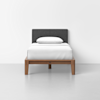 A single bed with a wooden frame, dark gray upholstered headboard, white bedding, and one white pillow, set against a plain white wall and light gray floor.