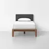 A single bed with a wooden frame, dark gray upholstered headboard, white bedding, and one white pillow, set against a plain white wall and light gray floor.