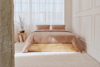 A neatly made bed with beige bedding and two pillows stands in a bright, minimalist bedroom with large windows, sheer curtains, and a wooden nightstand with a vase of flowers.