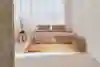 A neatly made bed with beige bedding and two pillows stands in a bright, minimalist bedroom with large windows, sheer curtains, and a wooden nightstand with a vase of flowers.