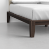 Close-up view of the corner of a wooden bed frame with dark brown legs and a white mattress in a minimal, modern bedroom setting with a light gray floor.
