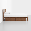 A wooden bed frame with a simple headboard, a white mattress, white bedding, and two storage drawers underneath, placed against a white wall on a light gray floor.