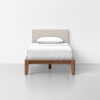 A single bed with a light beige upholstered headboard, white bedding, and a wooden frame, set against a plain white wall and a gray floor.