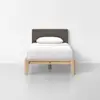 A twin bed with a light wood frame, brown fabric headboard, and white bedding with a single pillow, set against a plain white wall and gray floor.