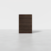 A modern, dark brown wooden chest of five drawers stands alone on a light gray floor against a plain white background.