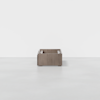 A small, box-shaped wooden table or storage unit with open sides and a minimalist design, placed in a white, empty room with a smooth floor.