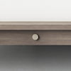 A close-up of a light wood bed frame with a round, cream-colored button that reads “SUPPLY MKT.” The background shows a light fabric and a gray floor.