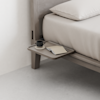 A minimalist bedside table attached to a wooden bed frame holds a black coffee cup on a saucer and an open notebook with a pen, next to neatly made beige bedding.