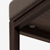 Close-up view of the corner of a wooden bench or table with a dark brown finish and a cushioned fabric top. A small round button or switch is visible underneath the surface.