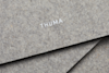 Close-up of textured gray felt material with the word THUMA printed in white, centered on the surface. Two overlapping pieces of felt are visible in the image.
