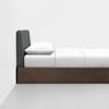 A neatly made single bed with a dark upholstered headboard, white bedding, and a wood frame, positioned against a white wall on a light gray floor.
