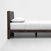 A minimalist bed with a dark wooden frame, white bedding, and a gray upholstered headboard, positioned against a plain white wall and floor.