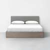 A modern bed with a light wooden frame, white bedding, two pillows, and a gray upholstered headboard, positioned against a plain white wall on a light gray floor.