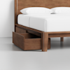 A wooden bed frame with white bedding and pillows features an open storage drawer built into the bed’s side, revealing extra space underneath.