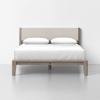 A modern wooden bed with a light upholstered headboard and white bedding, including two pillows, is centered against a plain white wall and sits on a light grey floor.