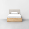 A single bed with a light wood frame, white bedding, and a light beige upholstered headboard, set against a plain white wall and a gray floor.