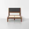 A wooden twin bed frame with a dark fabric padded headboard and slatted wooden base, set against a plain white wall and light gray floor.