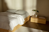 A minimalist bedroom with a wooden bed frame, white bedding, two pillows, and soft natural light. A low wooden nightstand holds a potted plant and two books, set on a hardwood floor with a light rug.