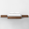 A minimalist bedroom setup with a wooden platform bed featuring white bedding and pillows, flanked by two matching wooden nightstands, all set against a plain white wall and gray floor.