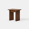A small, modern wooden side table with a rectangular top and two thick, straight legs on each side, connected by a horizontal support panel. The wood has a warm, medium-brown finish and the background is plain white.