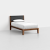 A single bed with a wooden frame, a dark upholstered headboard, and white bedding with one pillow, placed in a minimal, empty room with light gray flooring and white walls.