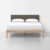 A modern wooden bed frame with a simple design, featuring a brown upholstered headboard, white bedding, and two white pillows, set against a plain white wall and floor background.