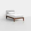 A single bed with a wooden frame, white bedding, a single pillow, and a light gray upholstered headboard, placed in a minimal, white room.