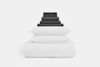 A neatly stacked bedding set with dark gray folded sheets and pillowcases on top, two white pillows in the middle, and a folded white comforter at the bottom, all against a plain white background.